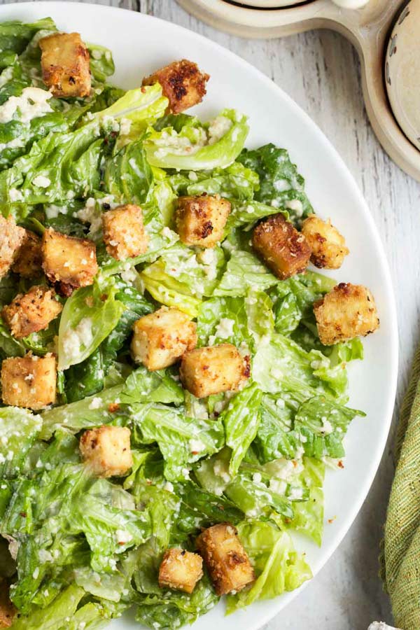 A top down close up shot of a white plate filled with vegan caesar salad with sesame tofu croutons layered on top.