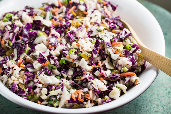 A white bowl of poppyseed coleslaw with a large wooden spoon.