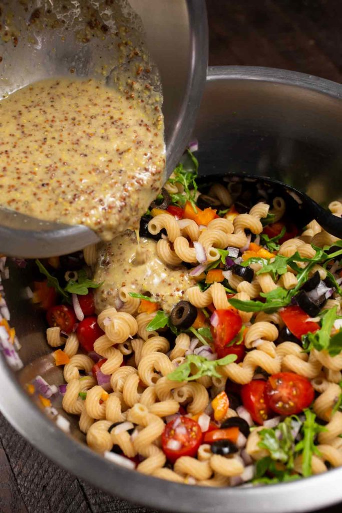 A bowl of colorful pasta salad with a bowl of sauce being poured on top of the pasta salad. 