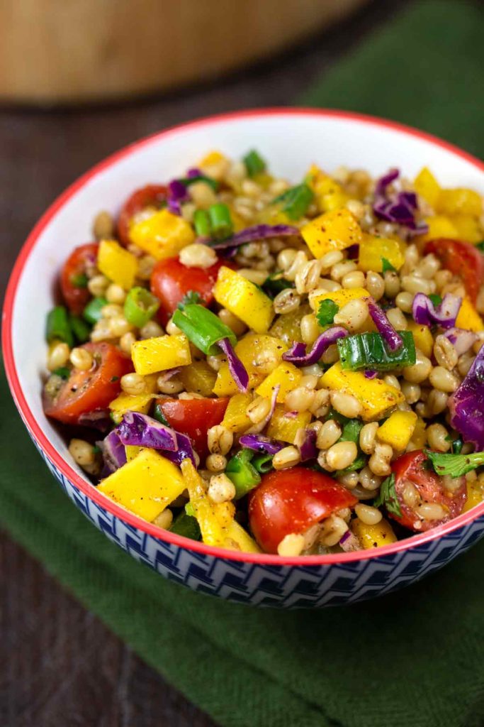 A closeup image of a bowl of wheat berry southwest salad, including chopped veggies such as yellow red pepper, mango, green onions, jalapeno, red cabbage and tomatoes. 