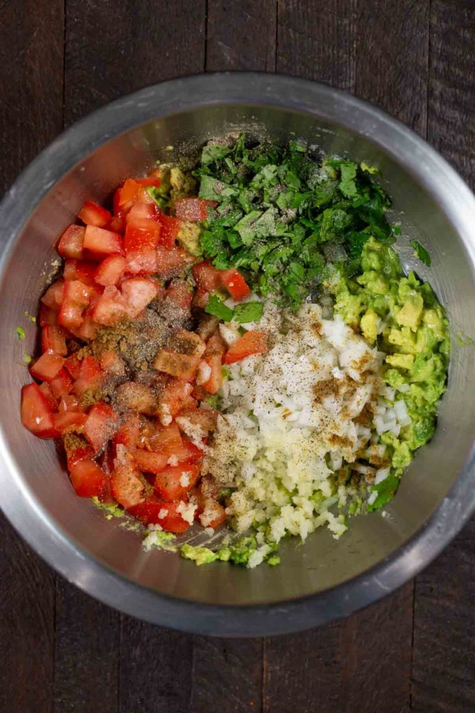 A stainless steel bowl filled with guacamole ingredients; diced tomatoes, chopped cilantro, diced white onion, pepper, salt, and lemon and lime juice. 