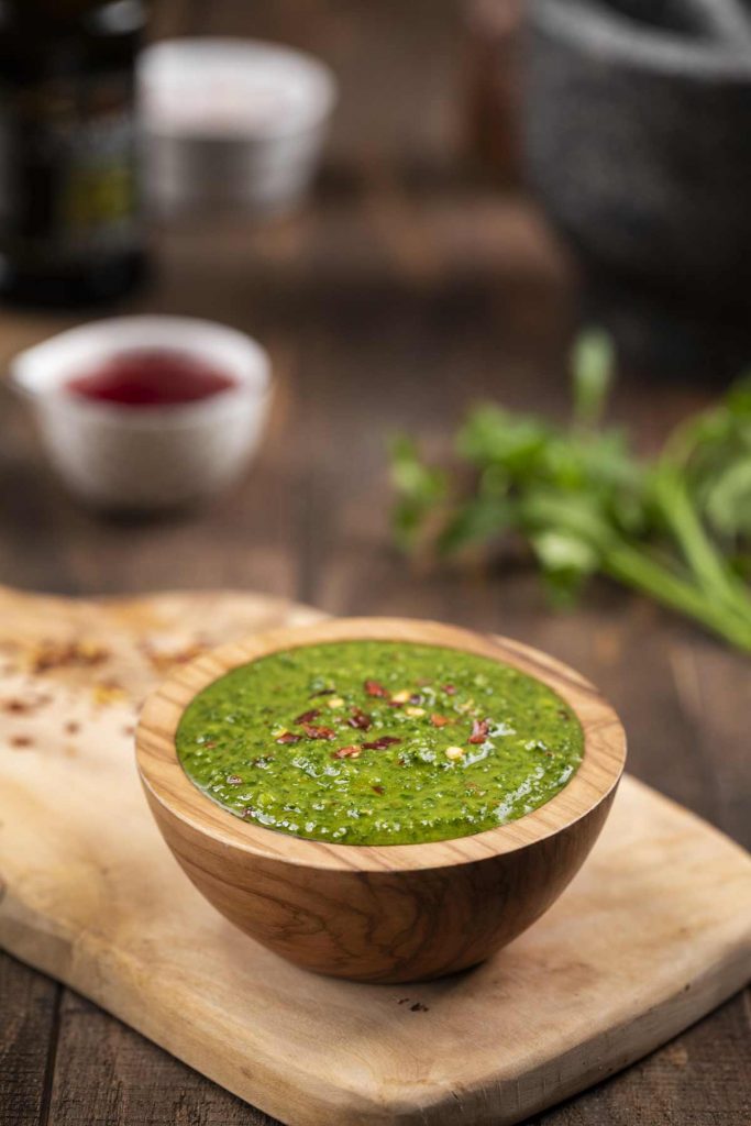 A wide shot showing a small cutting board with a small wooden bowl filled with chimichurri sauce, and a small white bowl filled with red wine vinegar and fresh parsley in the background.