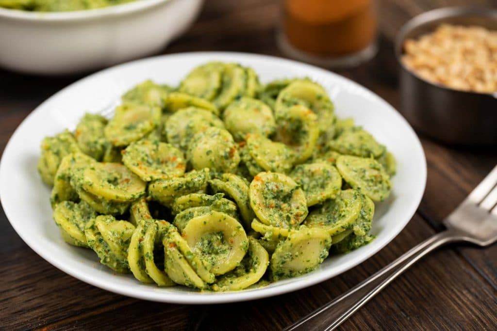 A white plate of pasta mixed with pesto sauce with a fork on the side.