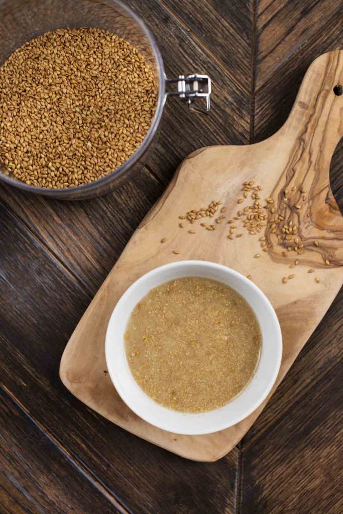 An overhead shot of a small cutting board with a bowl of flax egg on top, and a canister containing whole flax seeds on the side.
