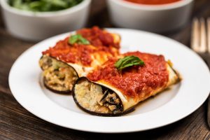 A white plate with 2 pieces of eggplant roll up casserole on top, with fresh basil and a side of sauce.