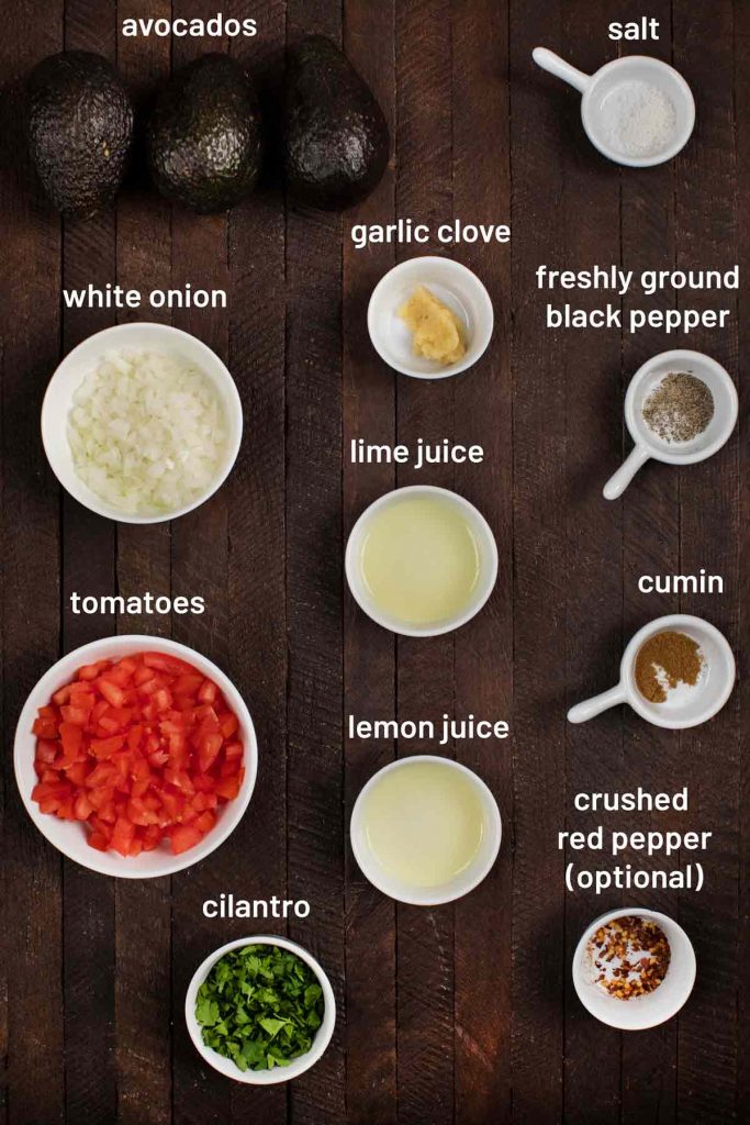 An overhead image of the ingredients for guacamole; avocados, salt, onion, garlic, black pepper, lime juice, lemon juice, cumin, tomatoes, red pepper, and cilantro.