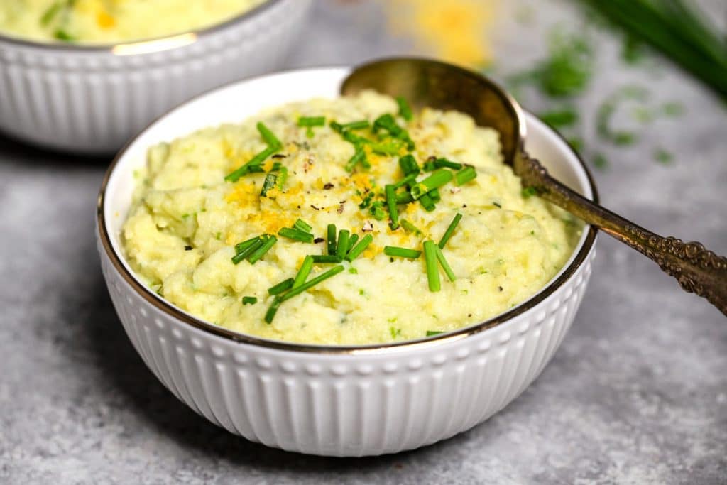 A closeup of a white bowl filled with vegan mashed cauliflower with chopped chives and nutritional yeast on top.