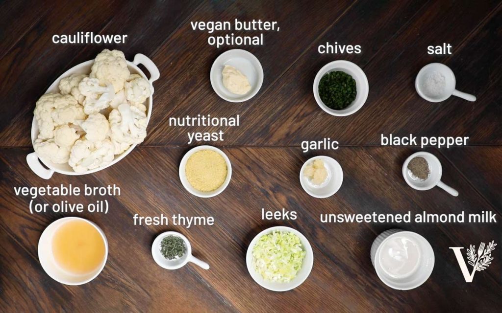The ingredients for mashed cauliflower, including cauliflower, butter, chives, nutritional yeast, broth, garlic, almond milk, and spices.