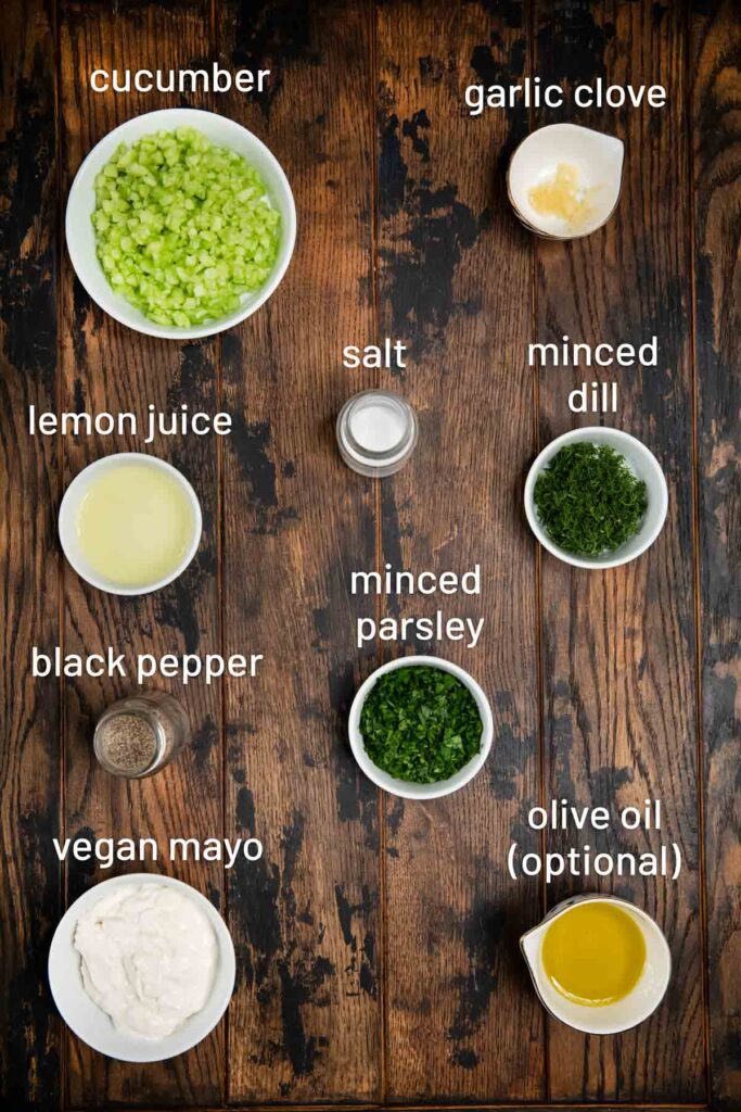 A wooden background with ingredients on top, such as cucumber, garlic, lemon juice, spices, dill, parsley, vegan mayo and olive oil.